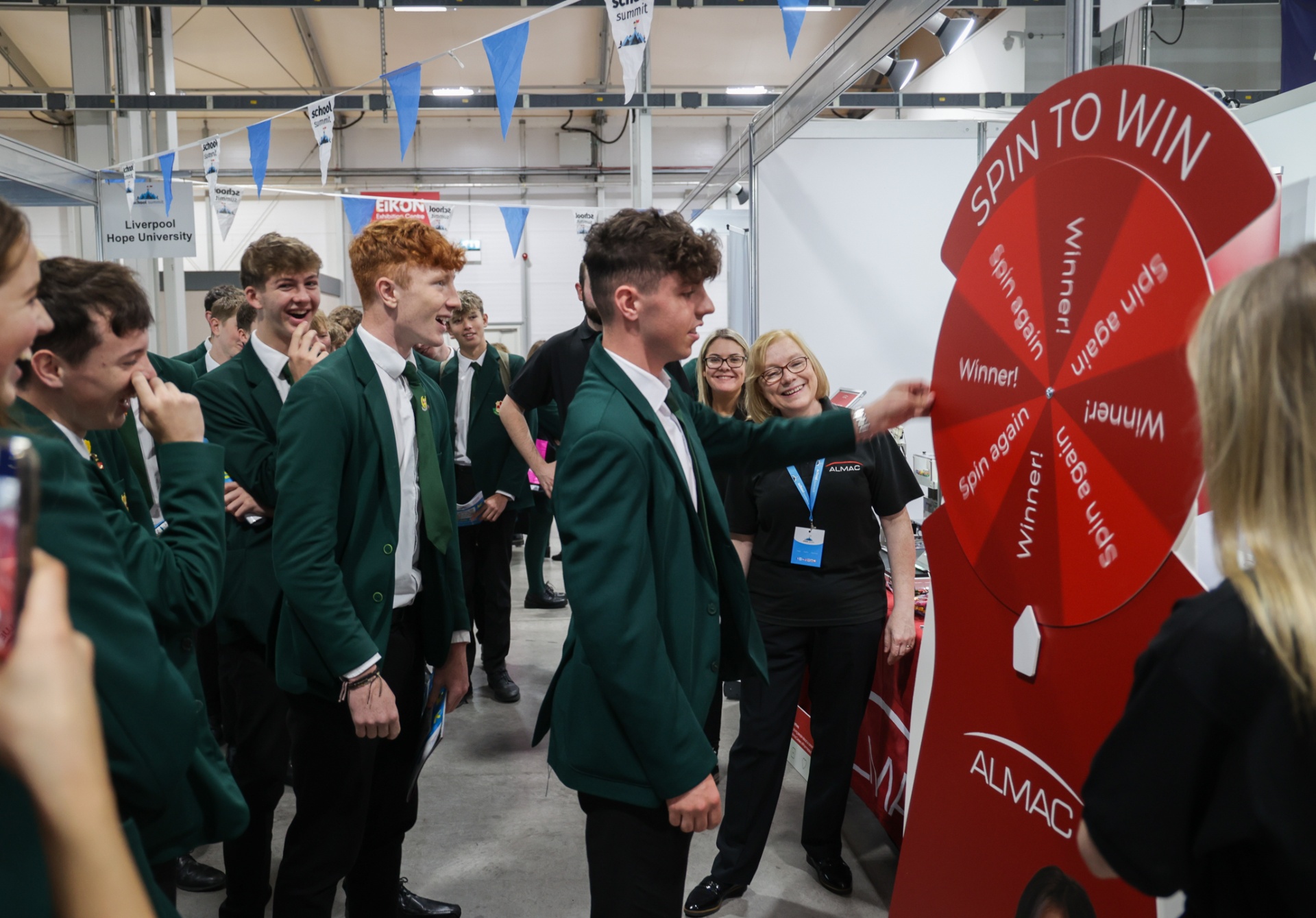 A crowd of pupils in green blazers gathered around the Almac ‘Spin to Win’ prize wheel at their School Summit Northern Ireland exhibitor stand, 2023.